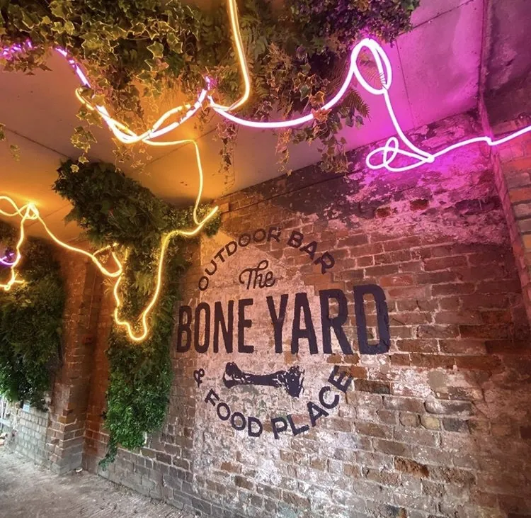 “The Boneyard — Outdoor Bar & Food Place” hand-painted in dark lettering on an exposed brick wall, lit by neon and trailing foliage.