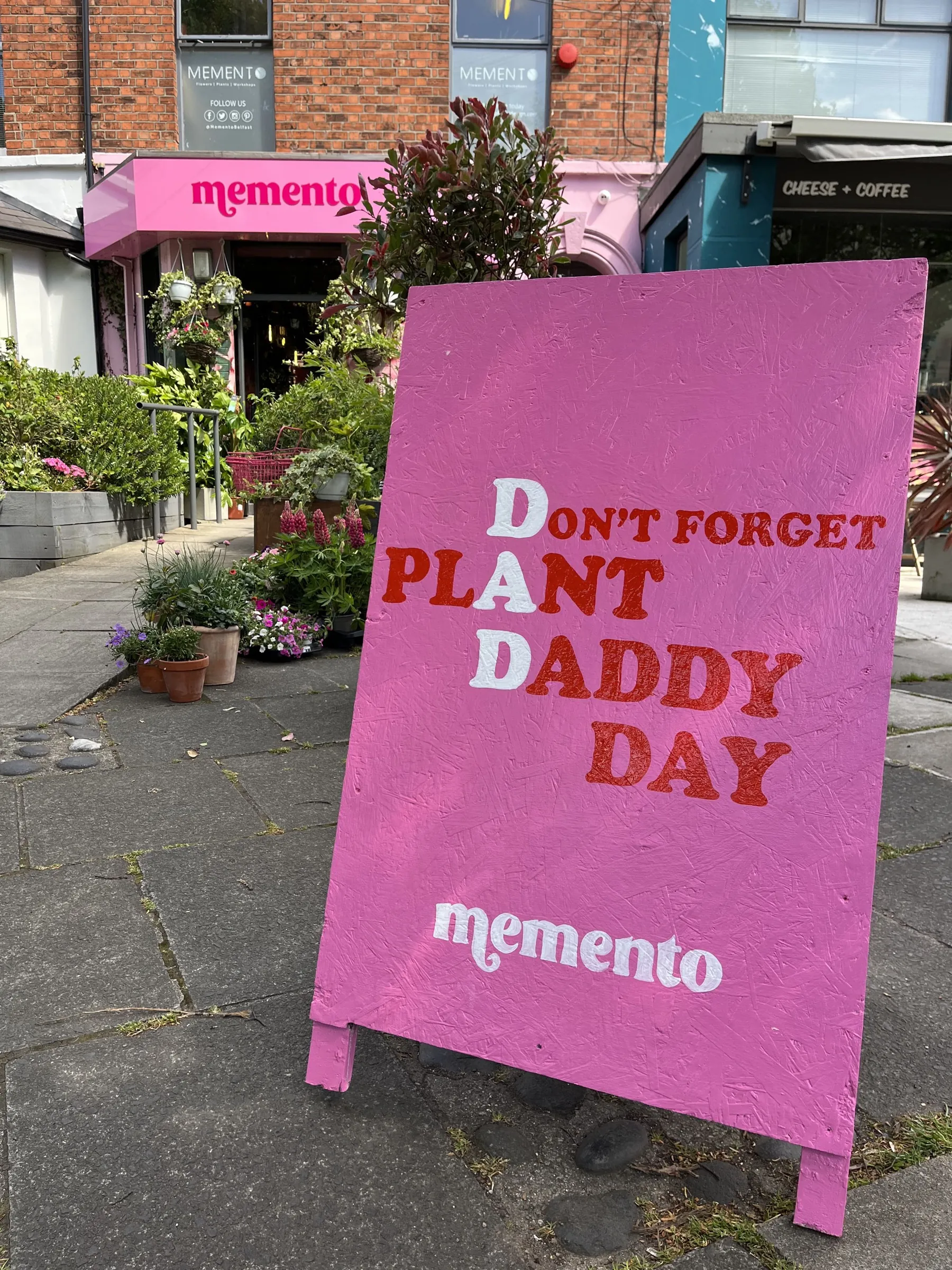 A pink A-board outside Memento, hand-painted with “Don’t forget Plant Daddy Day” in red and white lettering.
