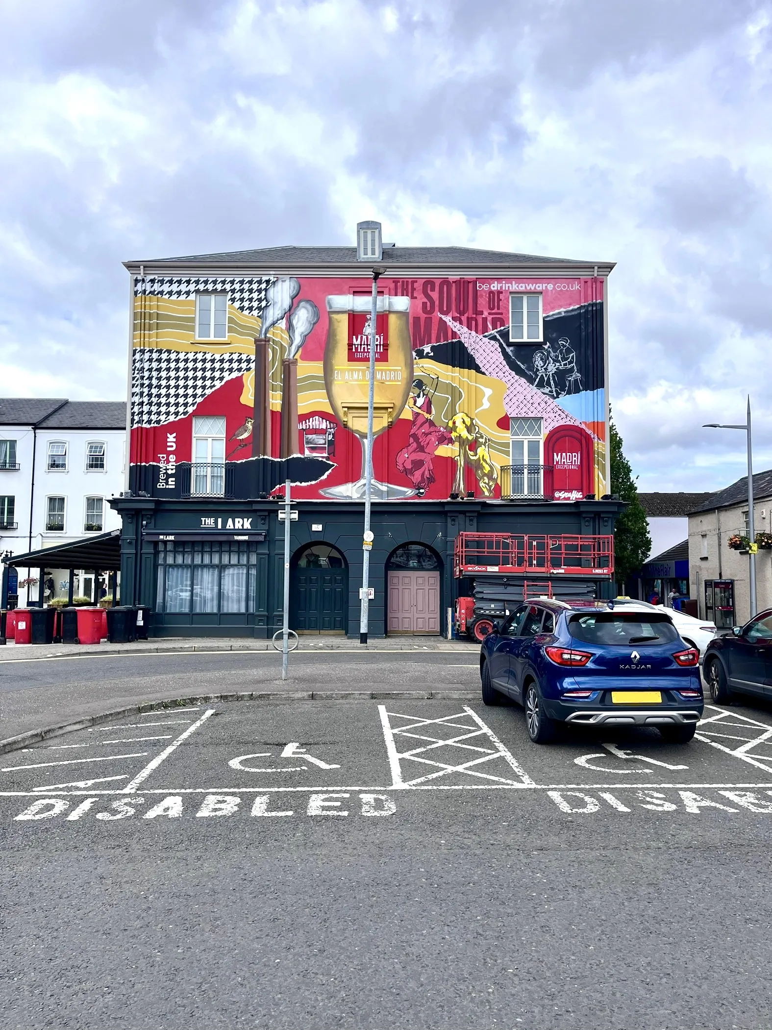A Madrí mural covering the upper floors of The Lark, a corner bar in Lisburn, in bold red, yellow and black shapes.