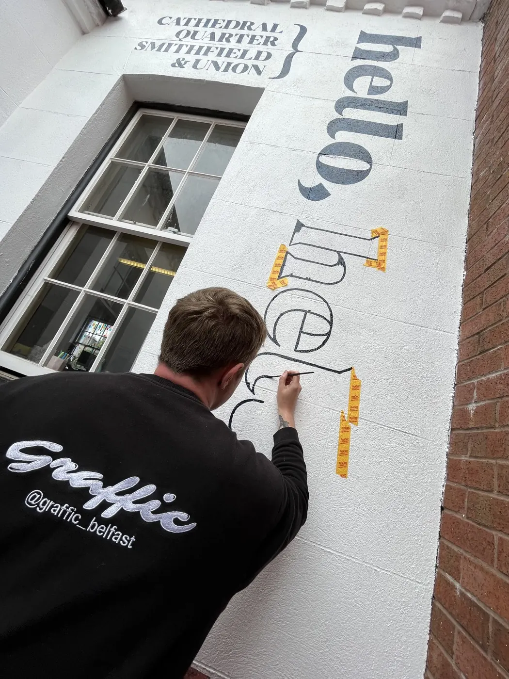Conor wears a black Graffic-branded hoodie with his back to the camera. He is painting the outline of a large, black letter onto a white wall. The wall has been masked with tape.