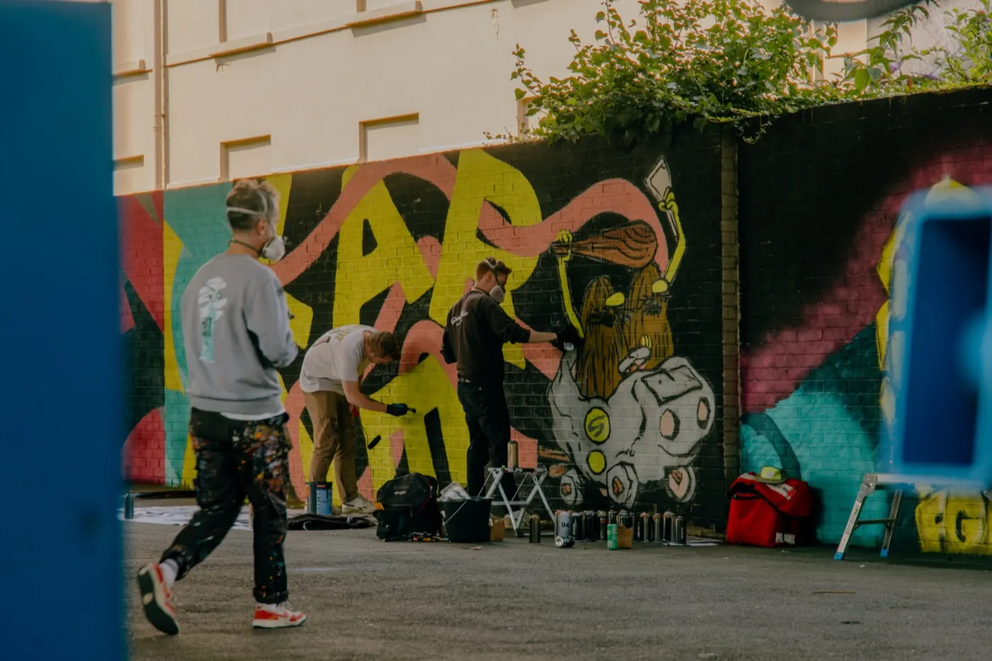 Conor and James of Graffic are painting a large, bright mural on a wall in Larne. Their spray cans and materials are at their feet, and the brothers are wearing ventilated masks. There is an unknown man in the foreground whose clothes are covered in paint, walking towards a mural he is working on.