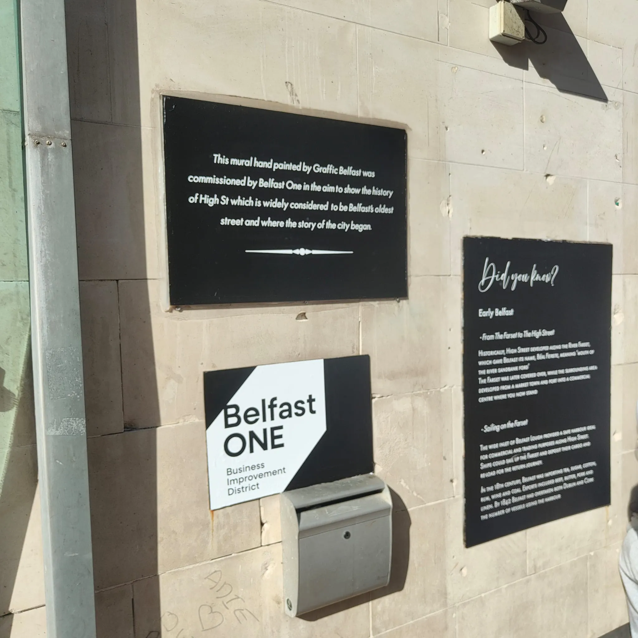 Black information panels and the Belfast One logo on the stone wall beside the mural, telling the story of High Street.