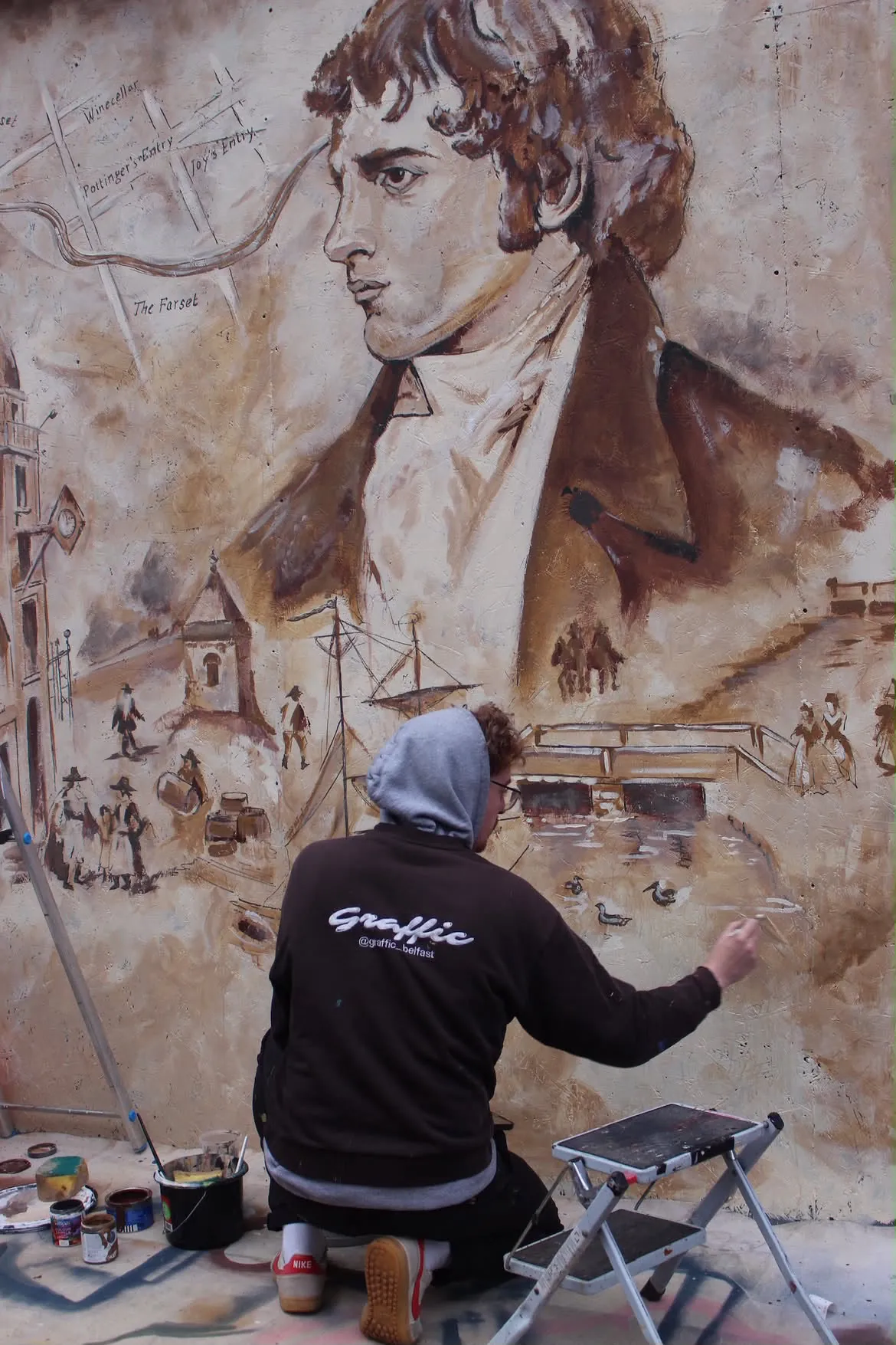 A Graffic painter kneeling to add detail to the old Belfast street scene below the mural’s portrait.