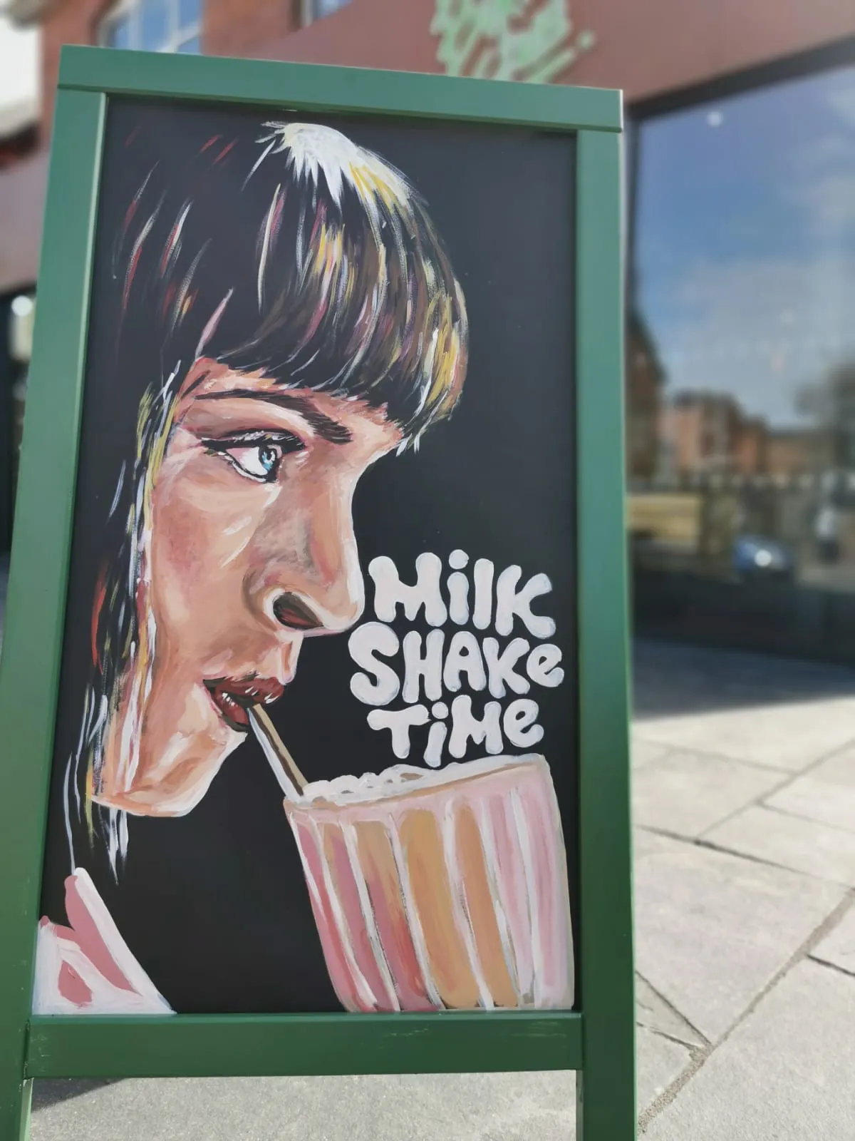 A green-framed A-board for The Burger Club, hand-painted with a woman drinking a milkshake and the words “Milk Shake Time”.