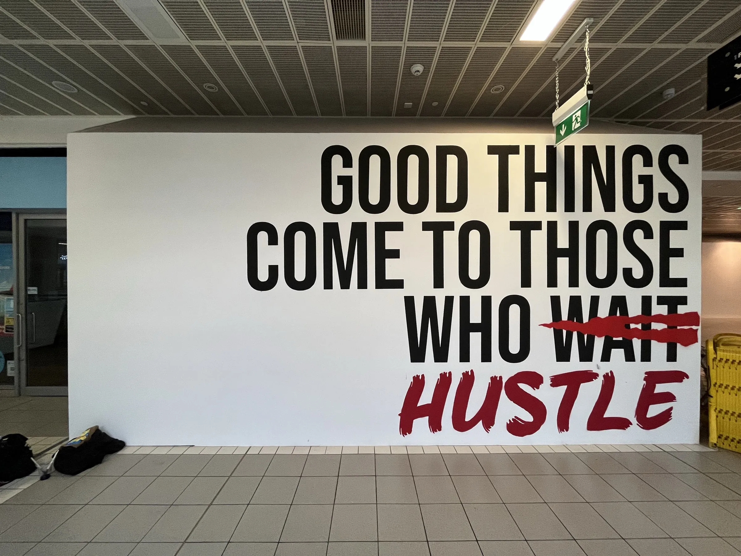 A CastleCourt hoarding painted with “Good things come to those who wait” in black capitals, with “wait” struck through and replaced by “hustle” in red script.