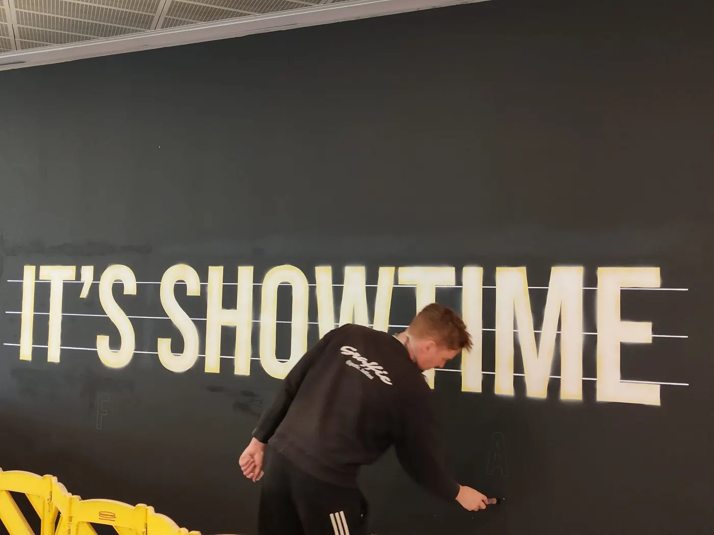 A Graffic painter filling in large cream capitals reading “It’s Showtime” on a dark hoarding, guide strings still pinned across the wall.