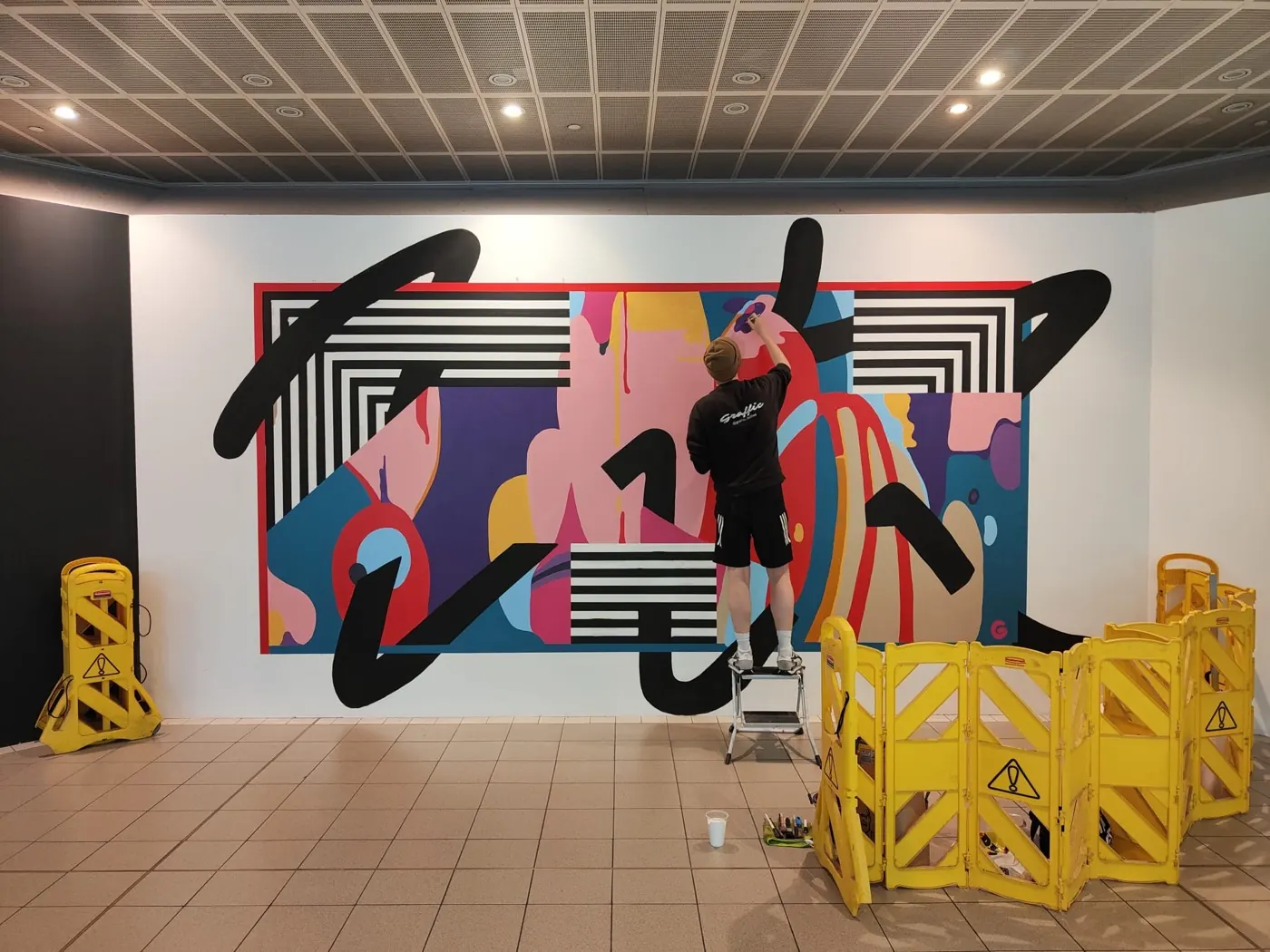 A painter on a step ladder laying sweeping black brush lettering over the finished abstract mural, yellow barriers around the work area.