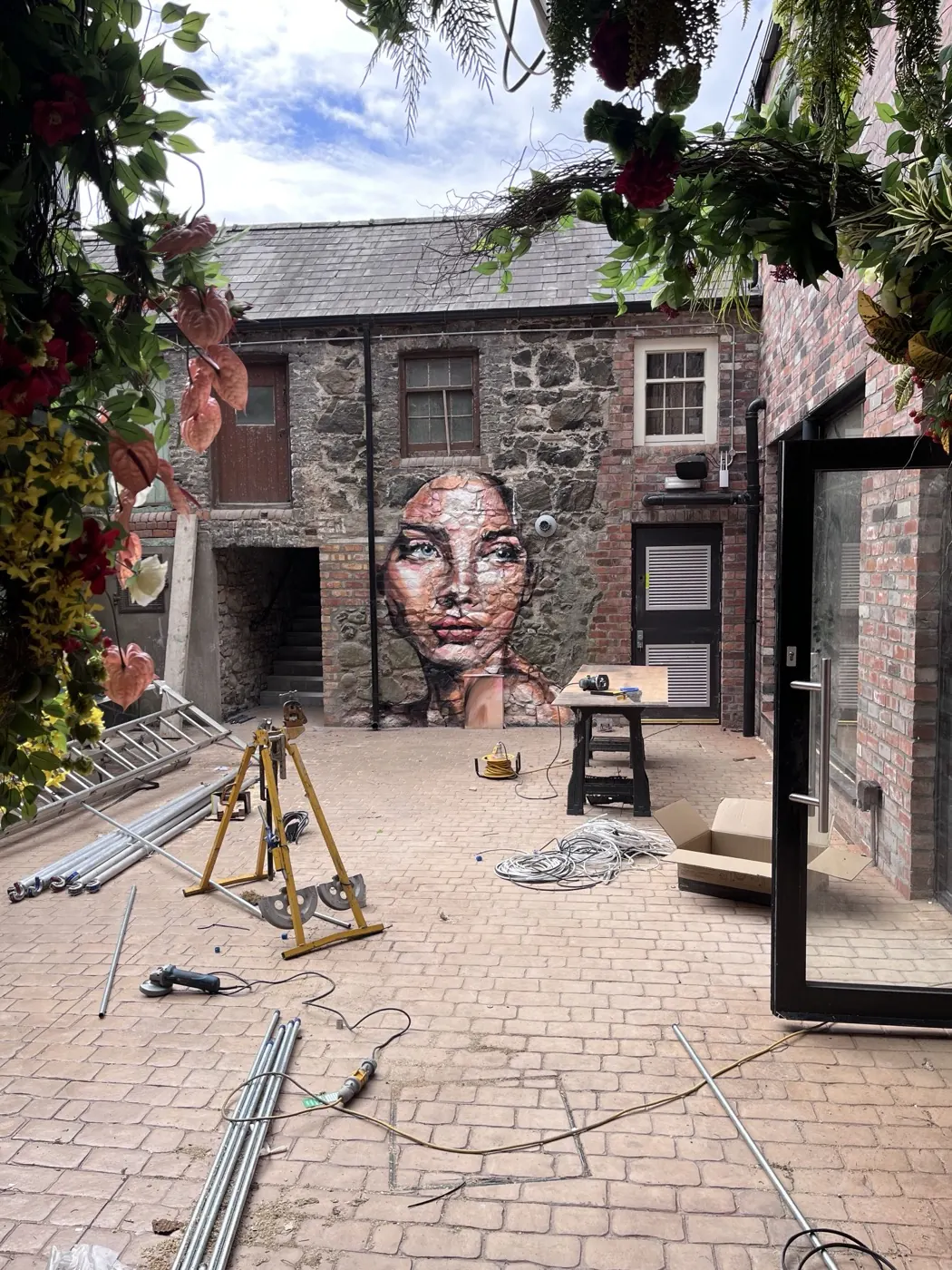 A woman’s face painted in soft tones across the farmhouse stone wall of the courtyard, with ladders and equipment still in the foreground.