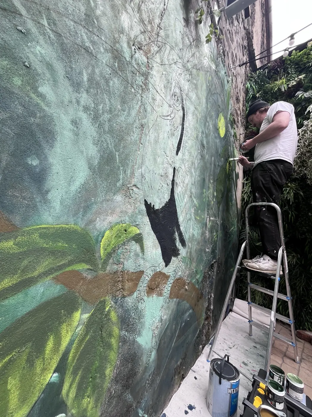 A painter on a step ladder working green foliage into the rainforest mural, paint tins lined up on the ground.