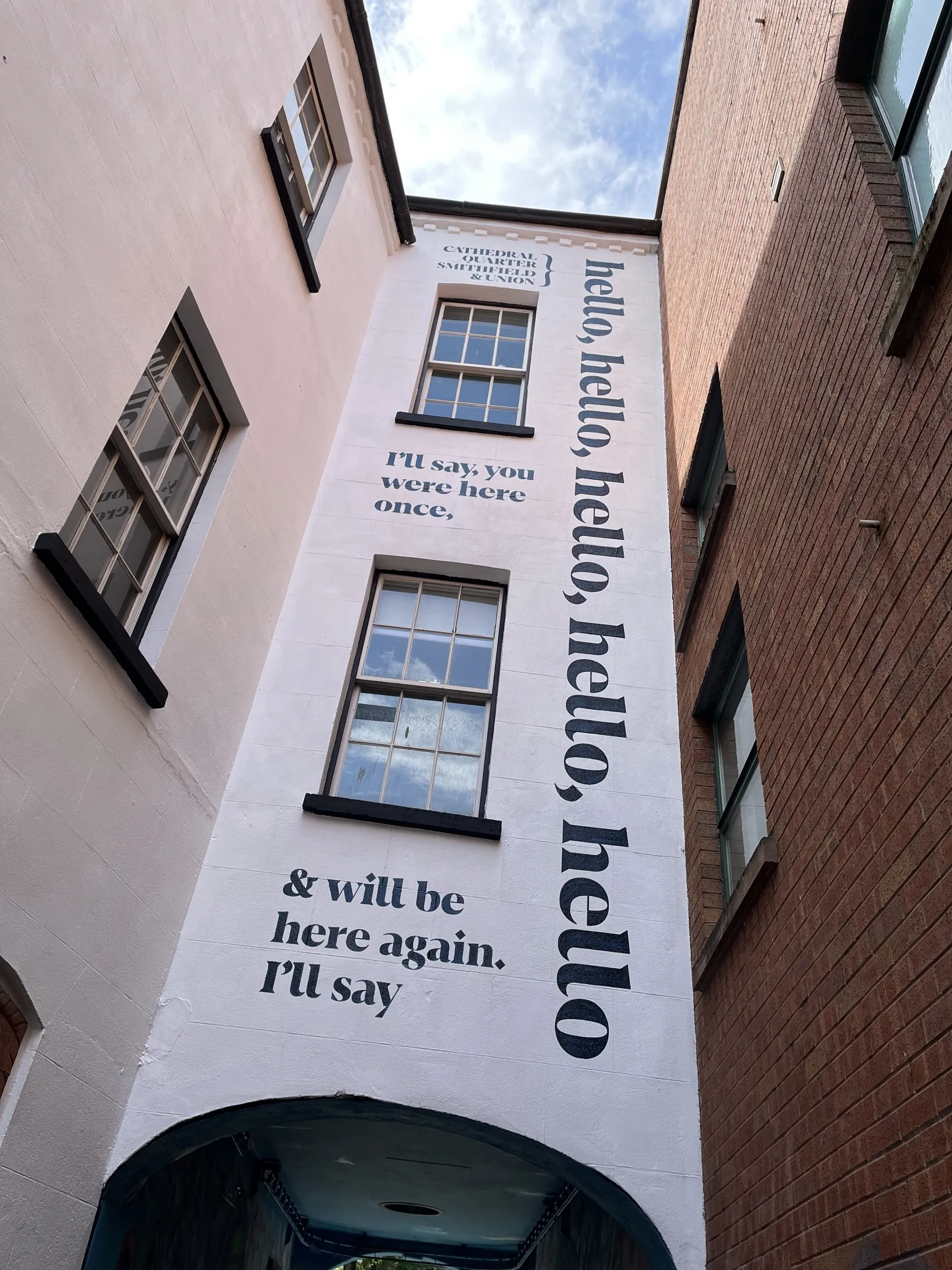 A tall white gable in Exchange Place hand-painted with a poem in black type, reading “I’ll say, you were here once, & will be here again. I’ll say hello, hello, hello.”