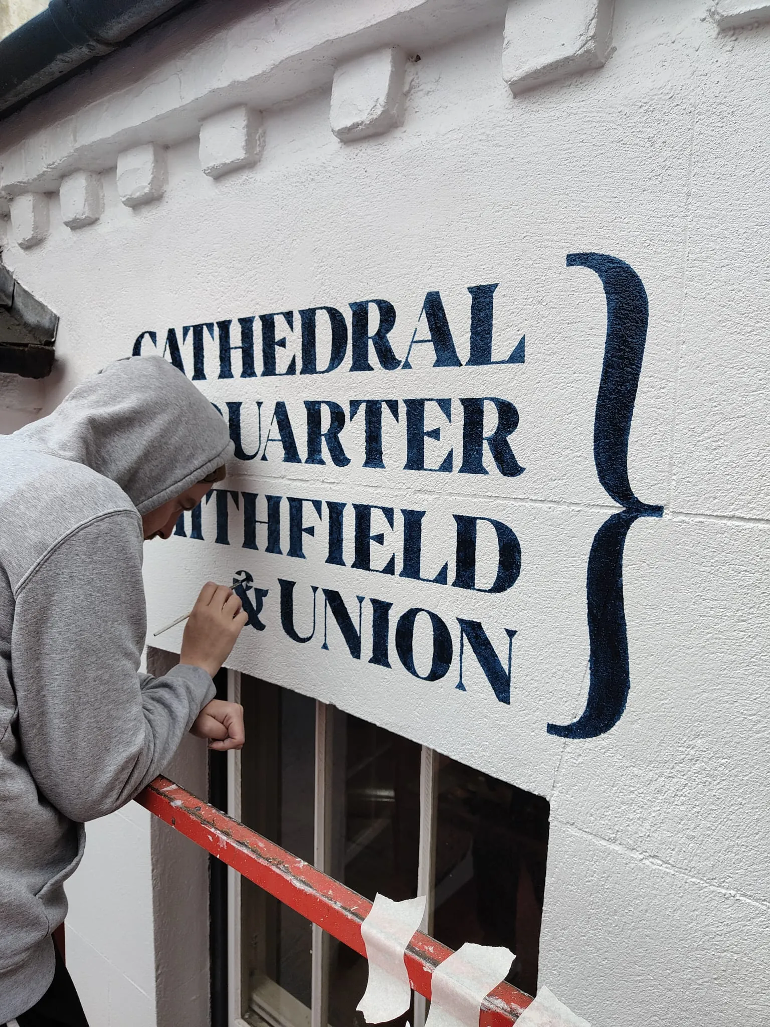 A painter leaning from a window to letter “Cathedral Quarter, Smithfield & Union” in navy on the white wall.