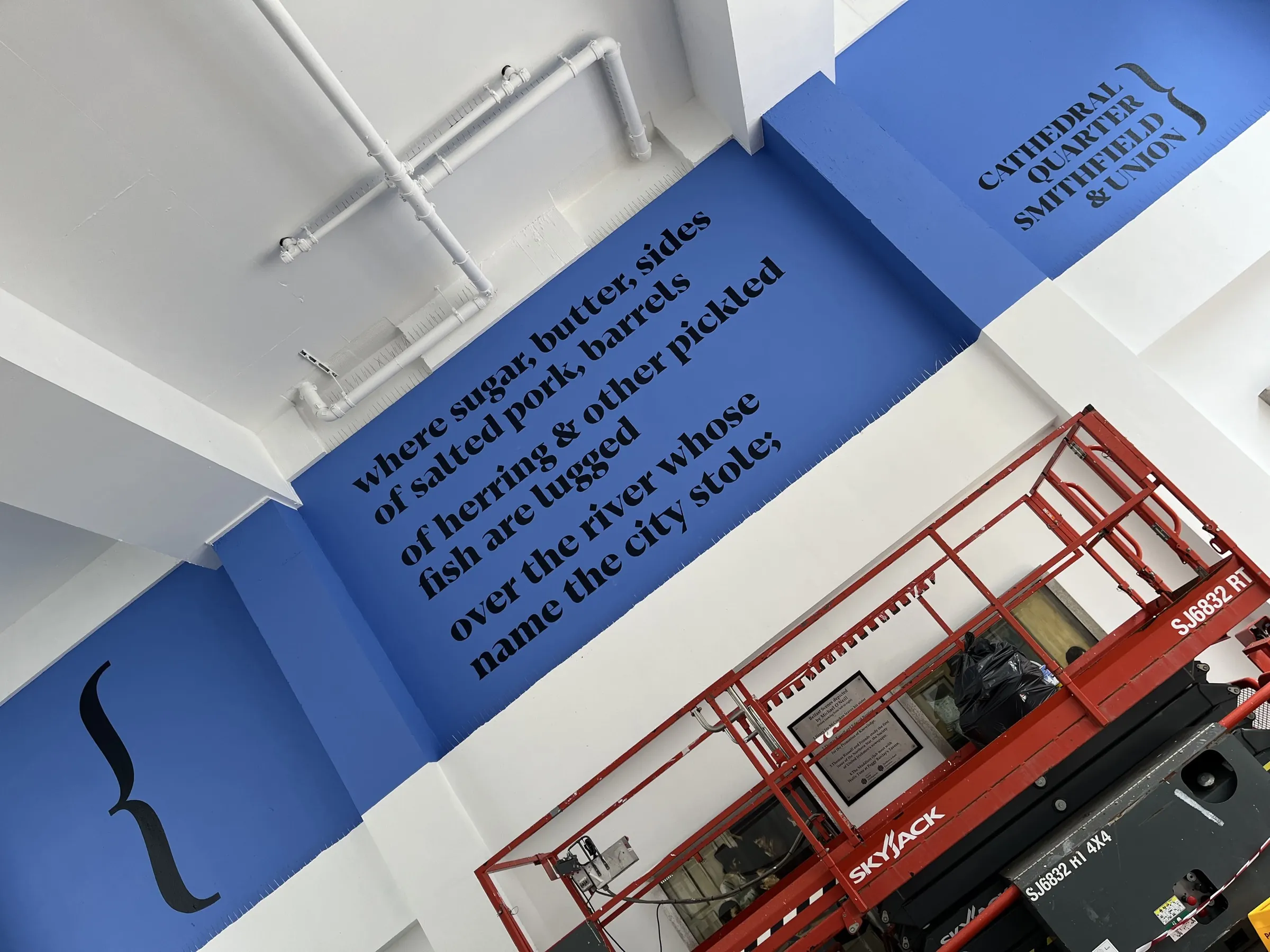 Lines of the poem painted in black type on a blue wall — “where sugar, butter, sides of salted pork, barrels of herring & other pickled fish are lugged over the river whose name the city stole;” — with a red scissor lift parked below.