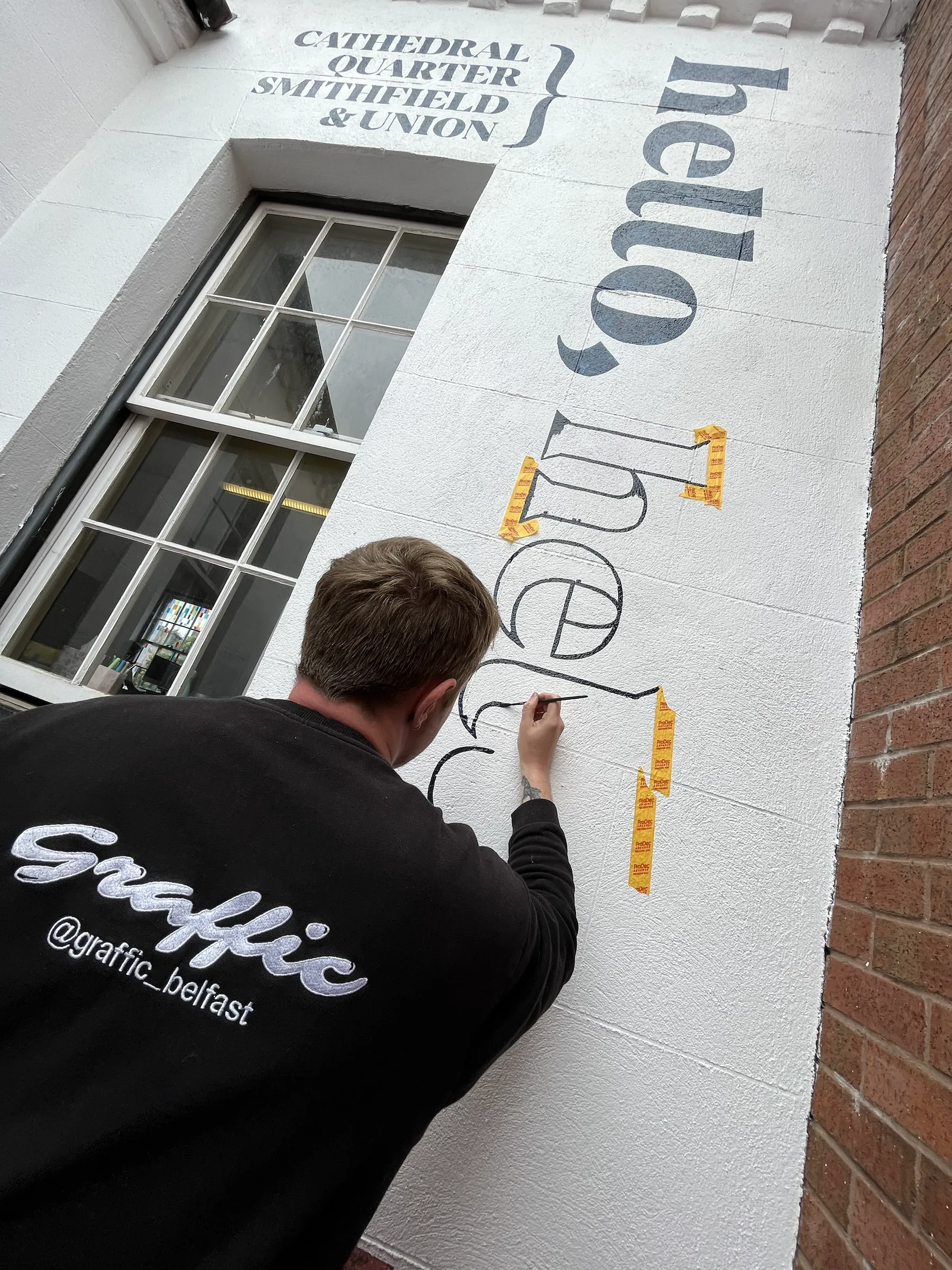 A Graffic painter lettering “hello” on the white gable beneath the “Cathedral Quarter, Smithfield & Union” credit, masking tape guides alongside.