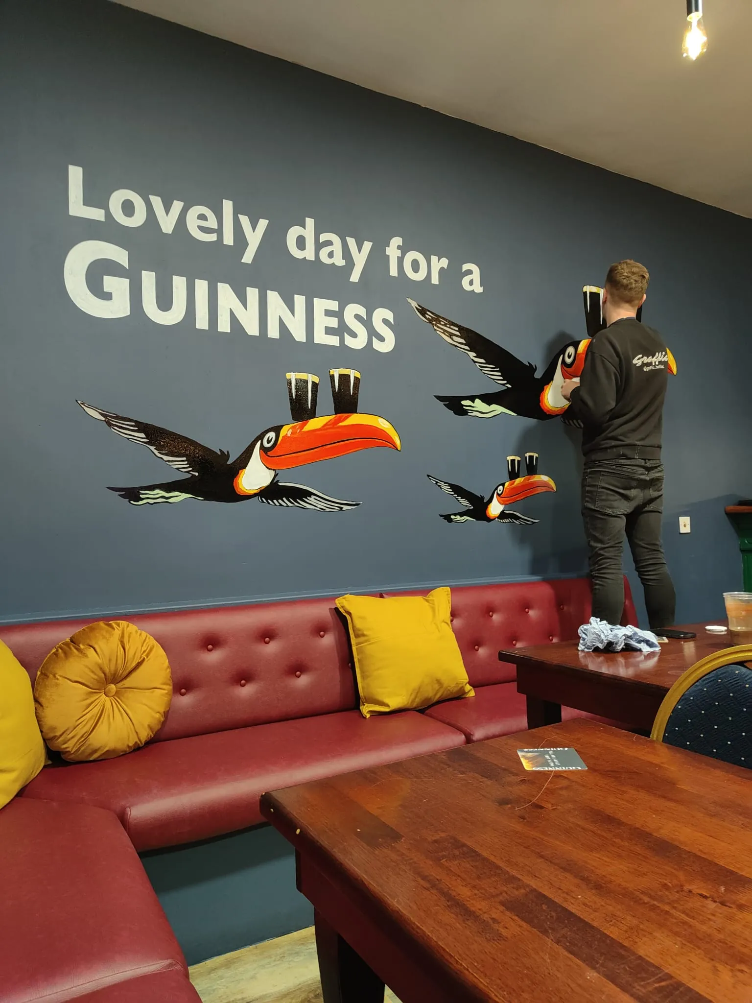 A painter adding a toucan to the “Lovely day for a Guinness” wall above red booth seating.