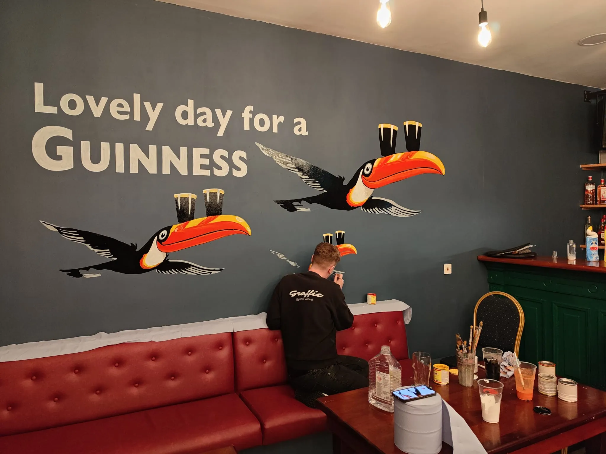 A Graffic painter kneeling on the red booth to add a small toucan to the “Lovely day for a Guinness” wall, paint pots and brushes on the table behind.