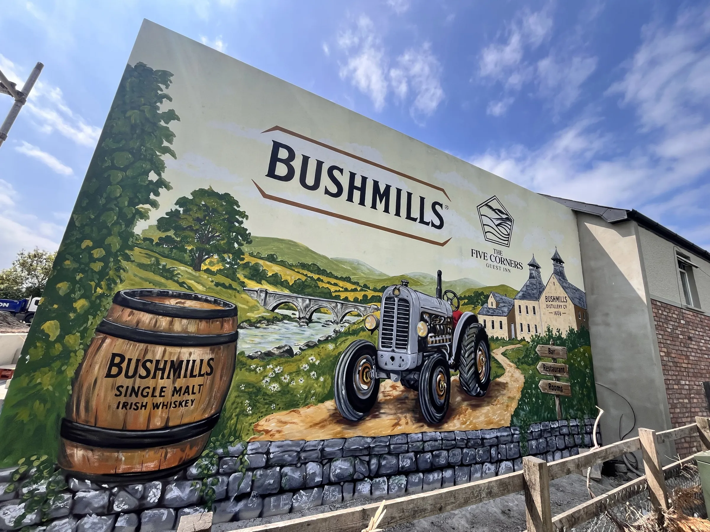 A hand-painted Bushmills mural on the gable of The Five Corners Inn, showing a grey tractor on a country road, a whiskey barrel and the Antrim countryside behind.