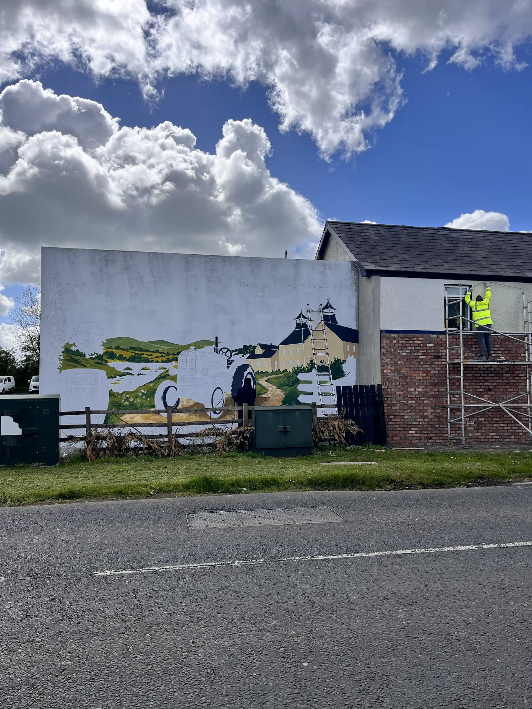 The mural half painted, a grey tractor and the distillery taking shape, with scaffolding at the gable’s end.