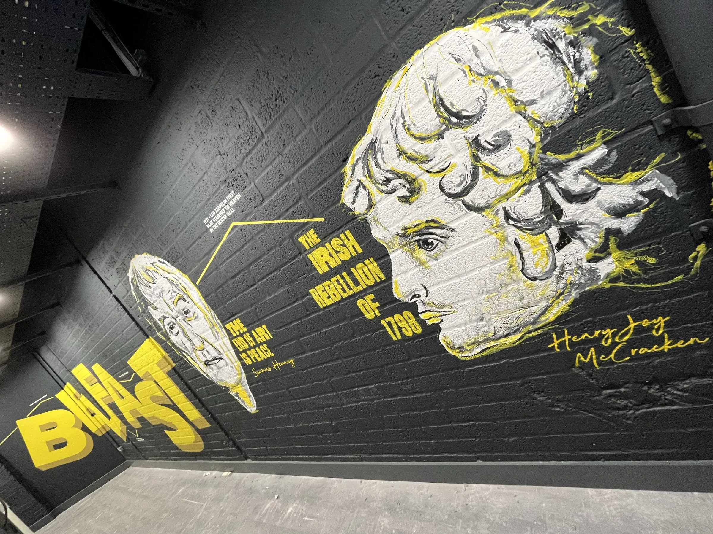A black brick wall painted with a yellow and white portrait of Henry Joy McCracken, captioned “The Irish Rebellion of 1798”, beside bold Belfast lettering.