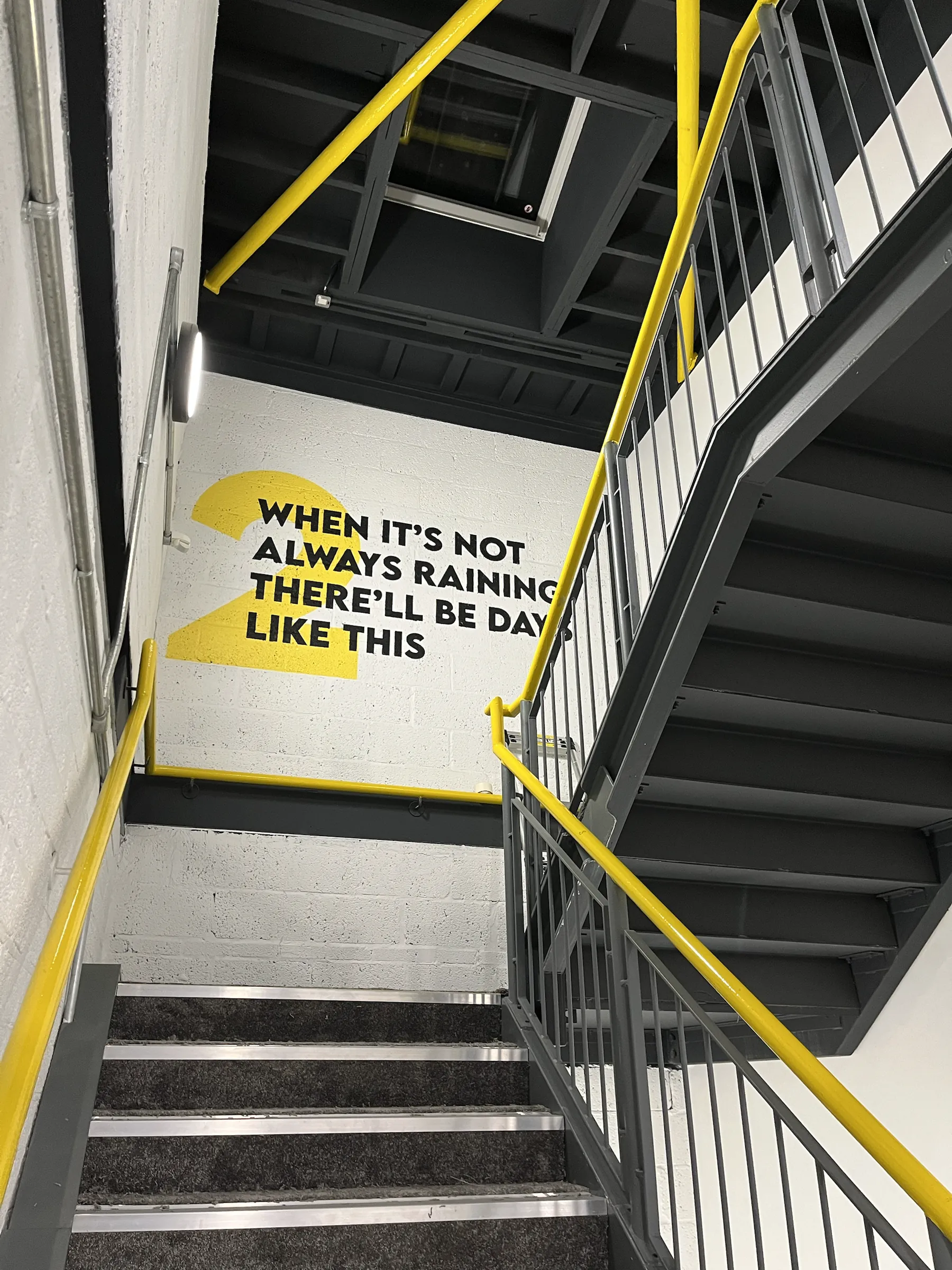 A stairwell wall painted with a yellow numeral and the line “When it’s not always raining there’ll be days like this”, beside yellow handrails.