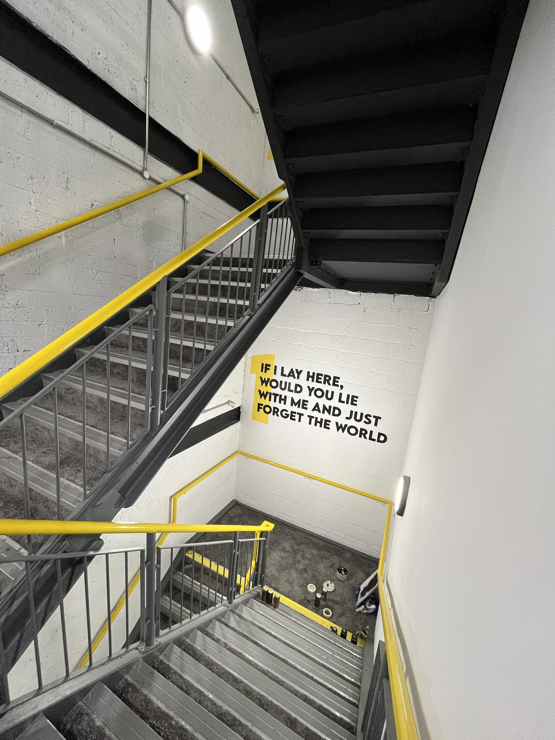 Looking down a stairwell to a wall painted “If I lay here, would you lie with me and just forget the world”.