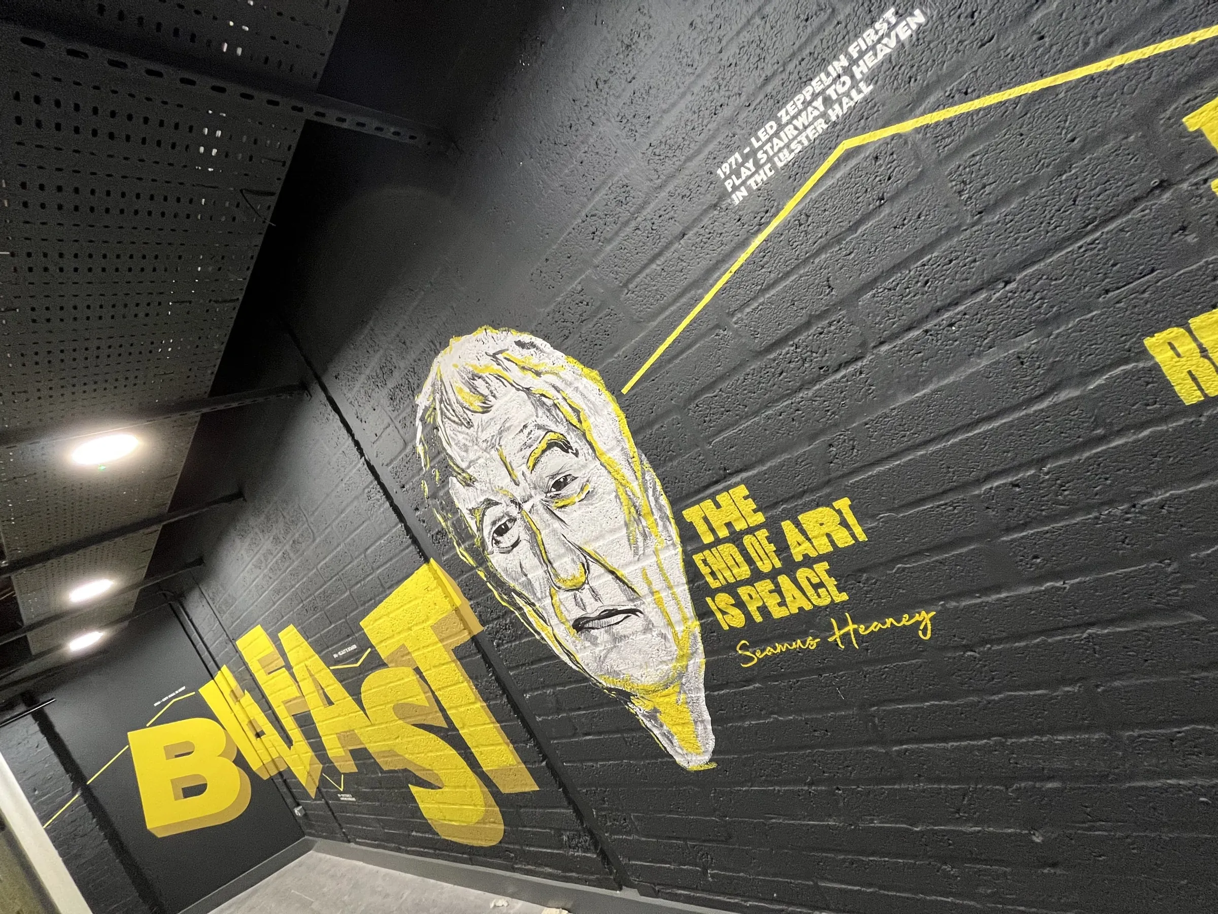 The black brick wall with its painted portrait and yellow “Belfast” lettering, seen at an angle.