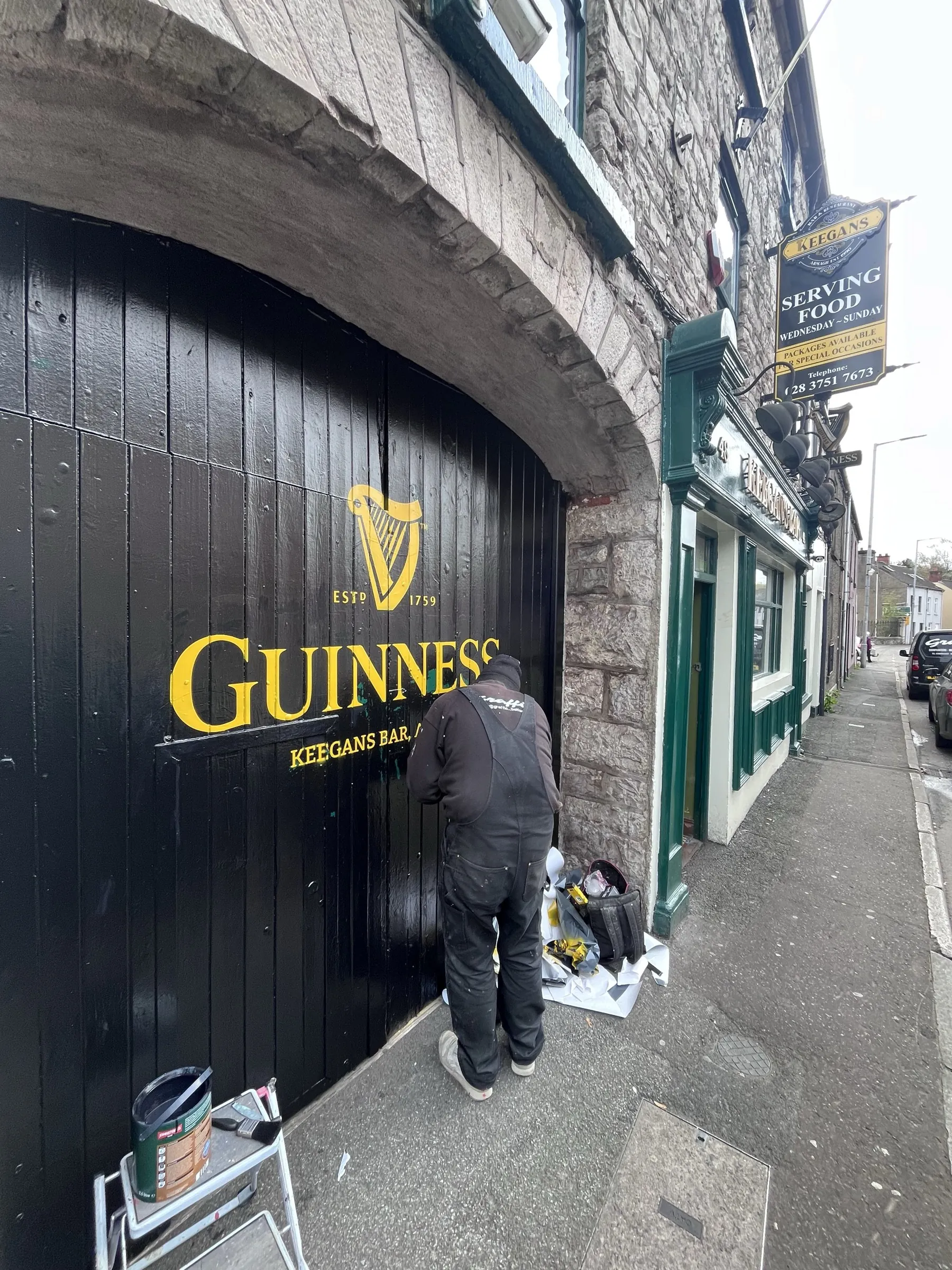 A signwriter hand-painting the gold Guinness harp and lettering onto the black timber shopfront of Keegan’s Bar.
