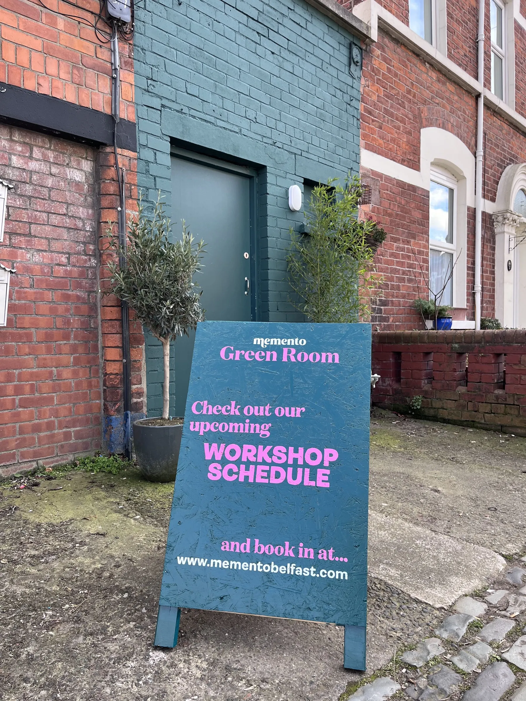 A teal Memento A-board outside a brick building, promoting the Green Room workshop schedule.
