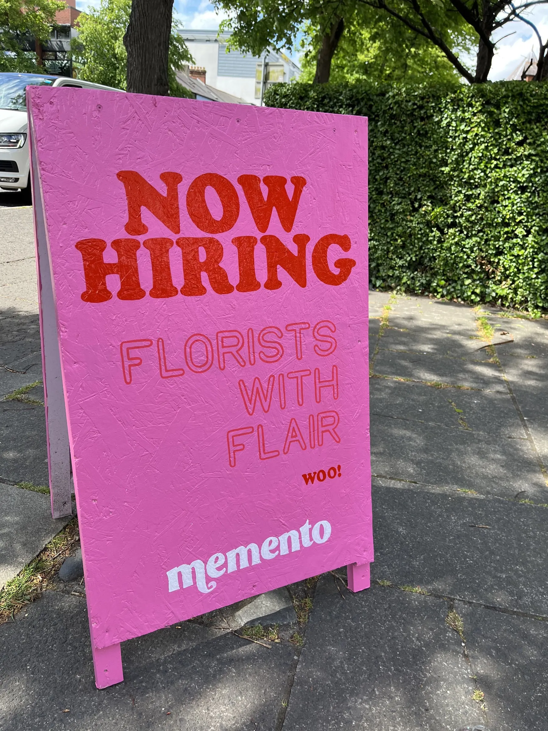 A pink Memento A-board painted with “Now hiring florists with flair” in red.