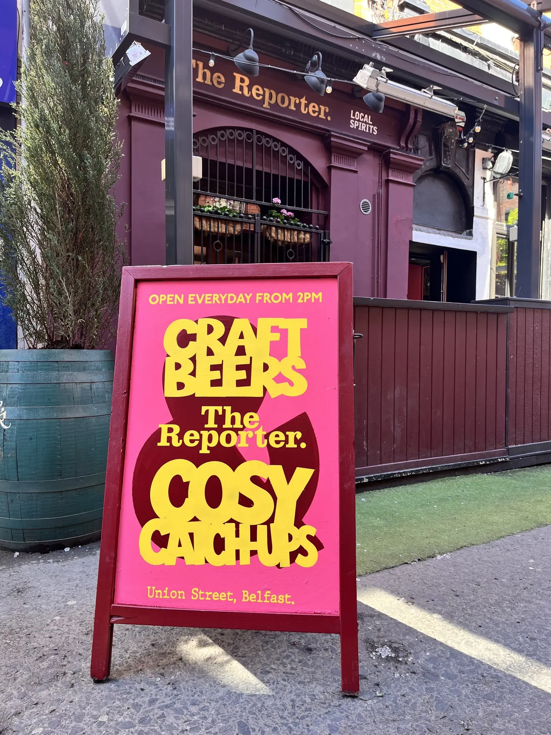 A pink A-board for The Reporter on Union Street, Belfast, hand-painted with “Craft beers & cosy catch-ups” in chunky yellow lettering over a maroon ampersand, standing on the pavement outside the pub.