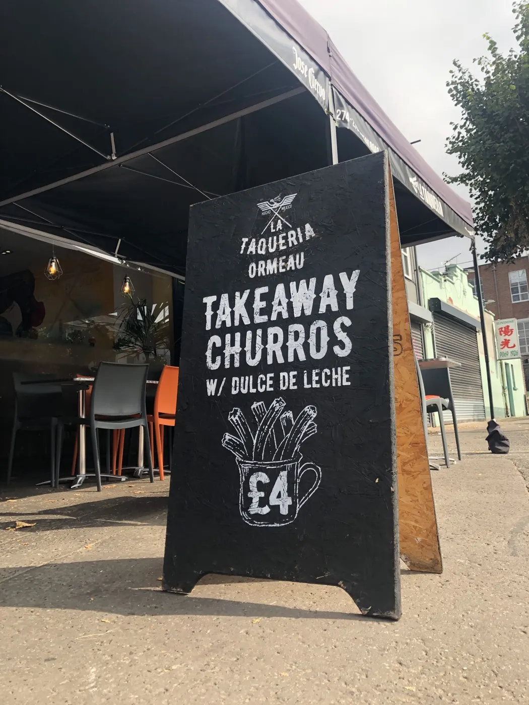 A black A-board outside Taqueria Ormeau, hand-painted with “Takeaway churros w/ dulce de leche, £4” and a drawing of churros in a cup.
