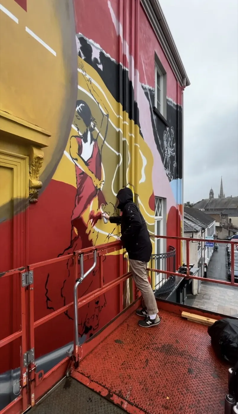 A painter on a raised platform working on a large red and yellow mural across the upper floors of a terraced building.