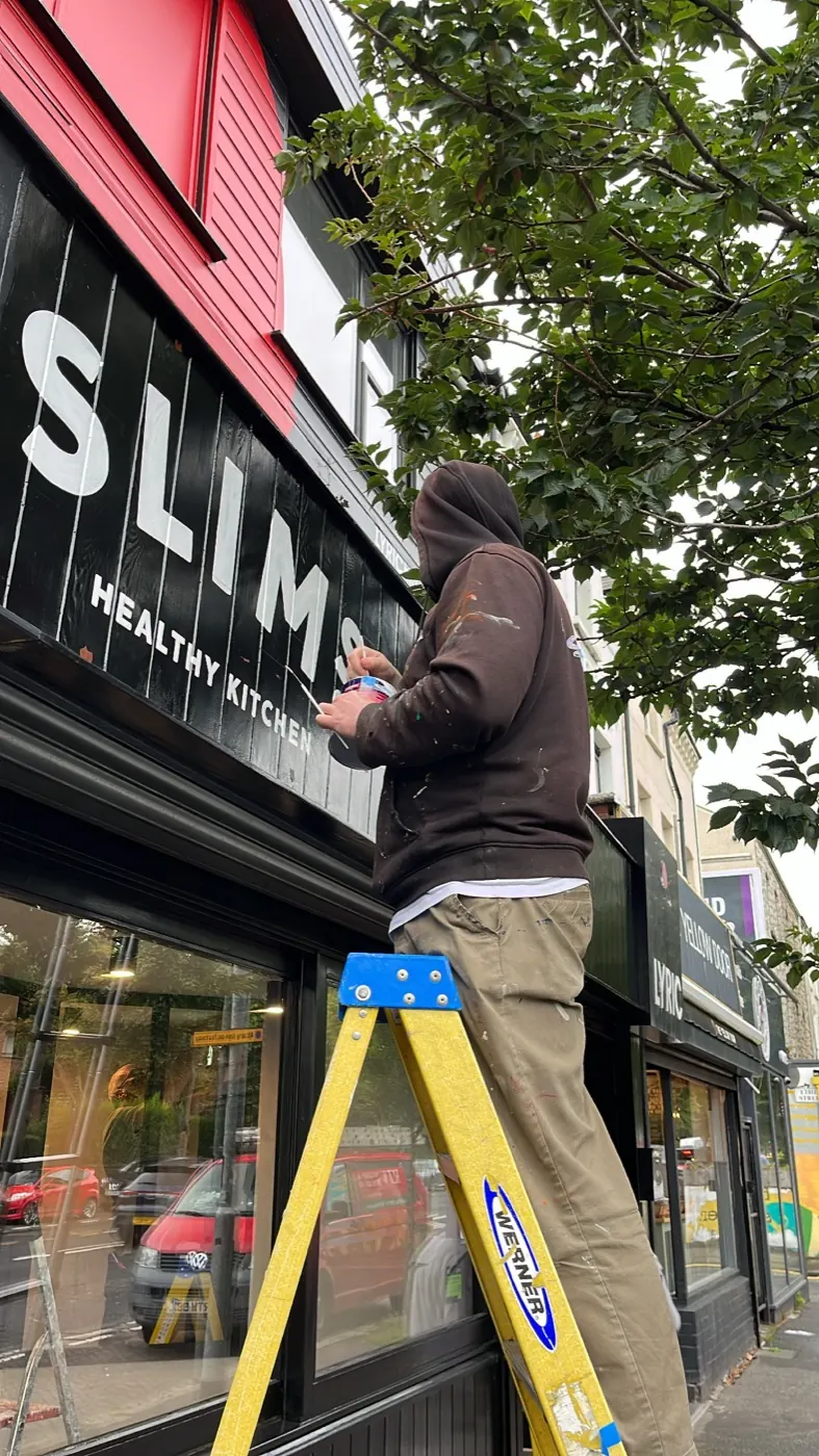 A signwriter on a step ladder hand-painting white lettering onto the dark shopfront fascia of Slim’s Healthy Kitchen.