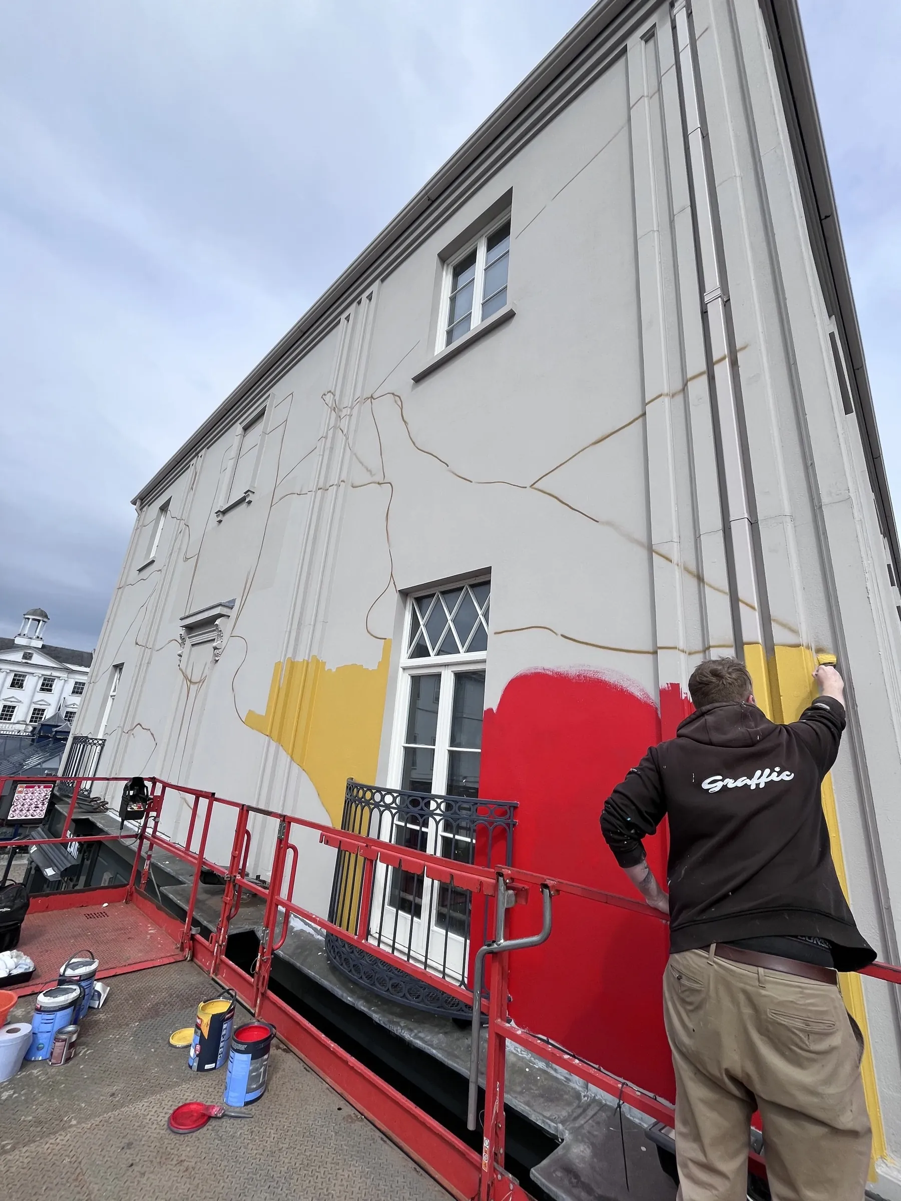 A painter on a cherry picker starting the red and yellow shapes of the Madrí mural on The Lark’s white wall.