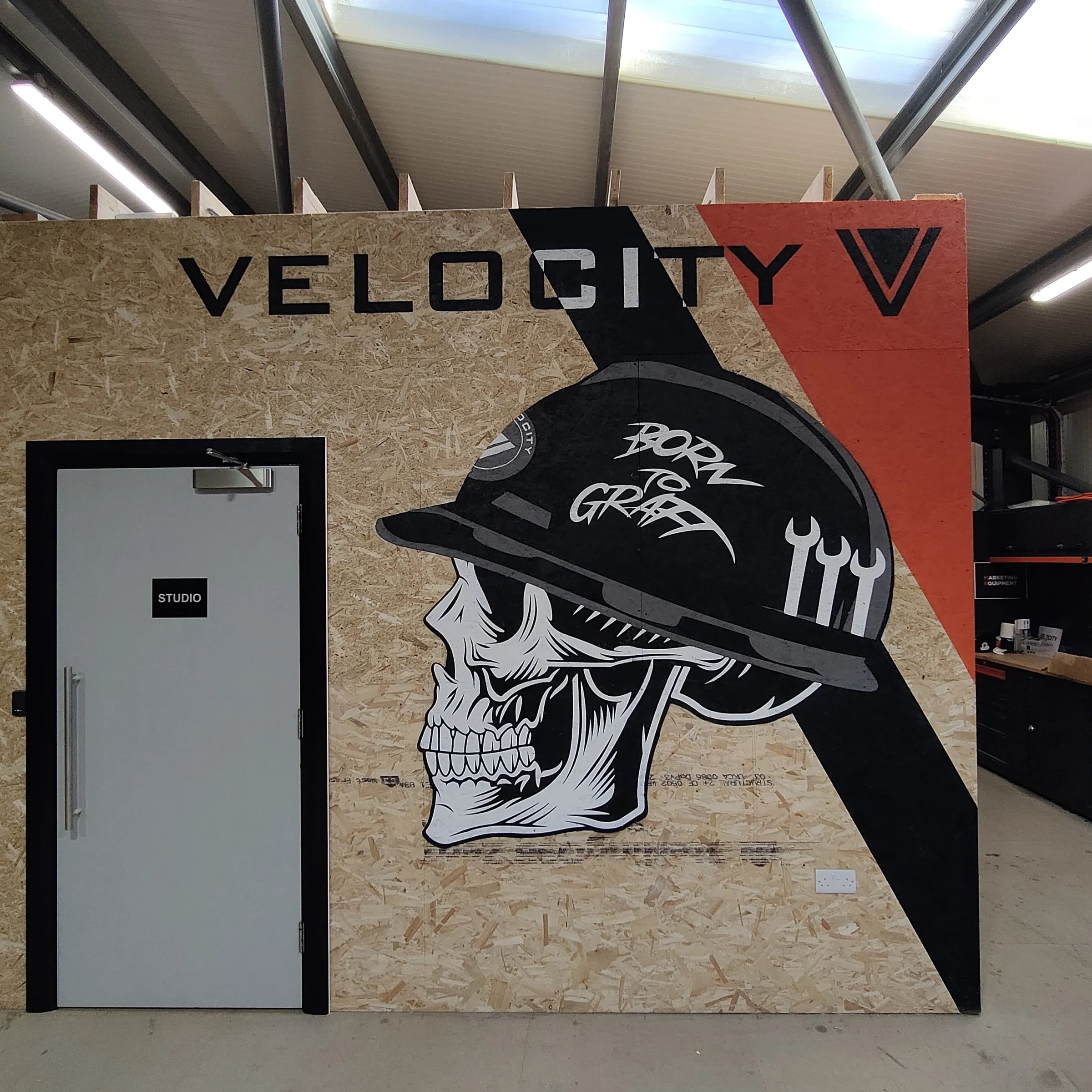 A hand-painted Velocity sign on a plywood wall: white block capitals beside a monochrome skull in a hard hat marked “Born to Graft”, over a black and orange chevron.