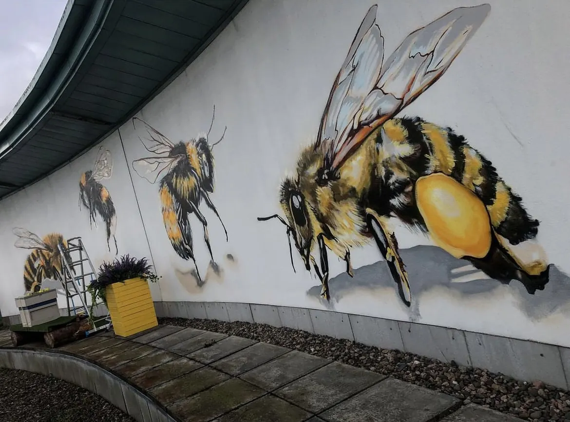 Three oversized bees — a honeybee and two bumblebees — hand-painted along the curved white wall at Victoria Square.