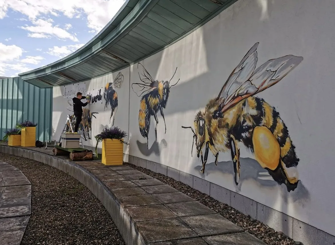 The curved Victoria Square wall with its row of hand-painted bees, a painter working at the far end beside yellow planters.