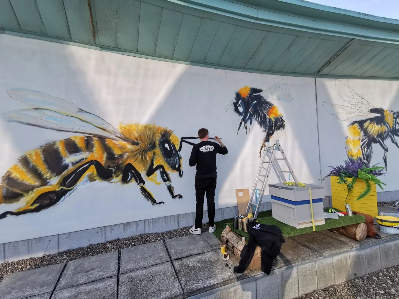 A painter in a Graffic hoodie adding detail to a bumblebee, the finished 8ft honeybee filling the wall to his left.