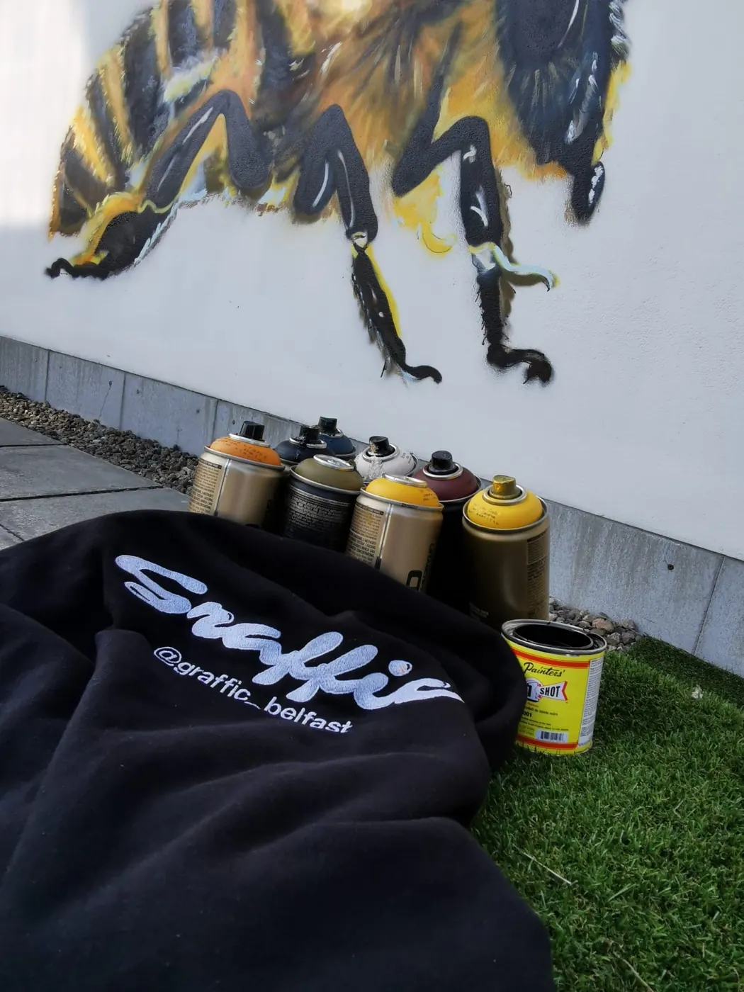 A Graffic hoodie and a row of spray cans on the ground in front of the finished honeybee mural.