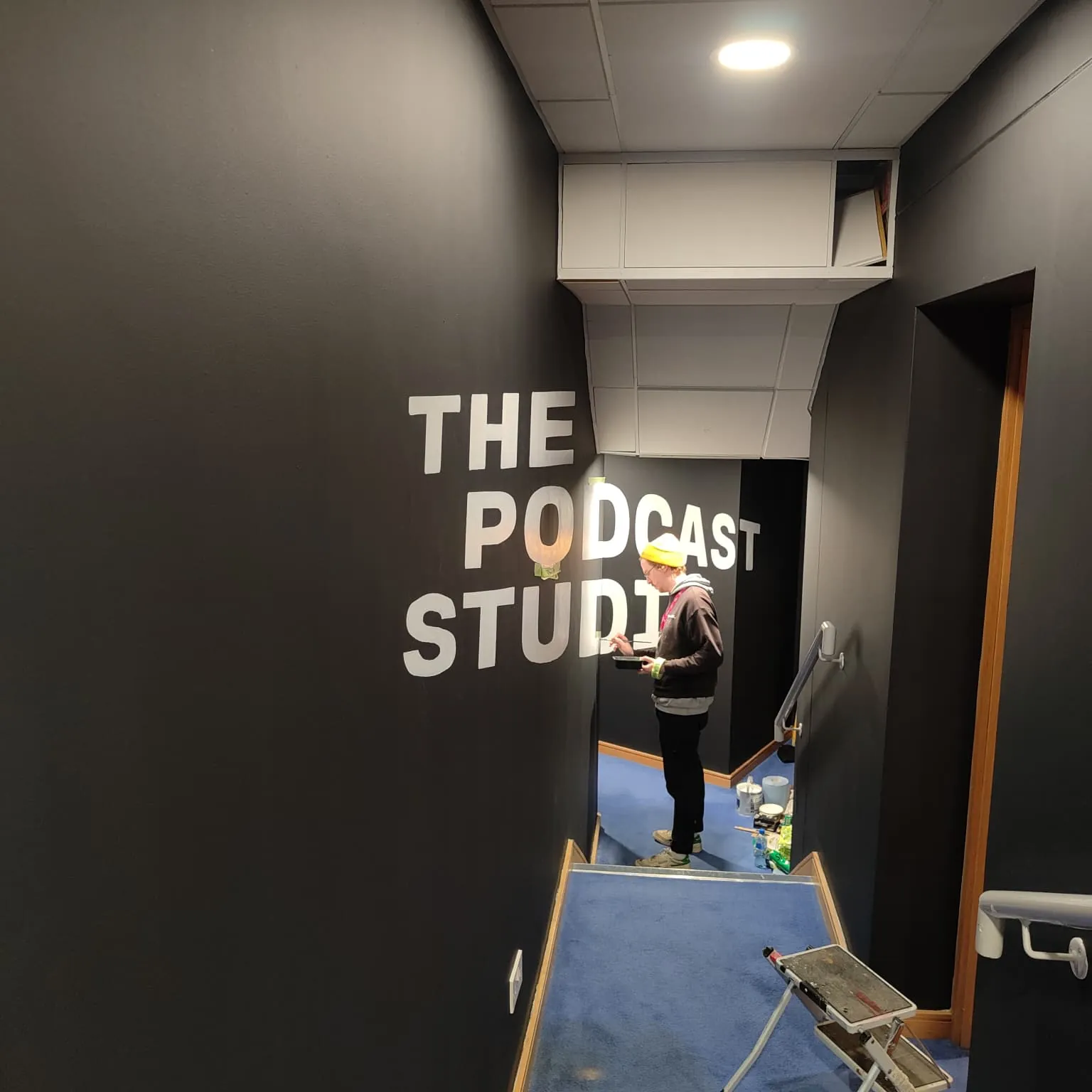 A painter at the far end of a narrow black corridor, lettering “The Podcast Studio” in white.