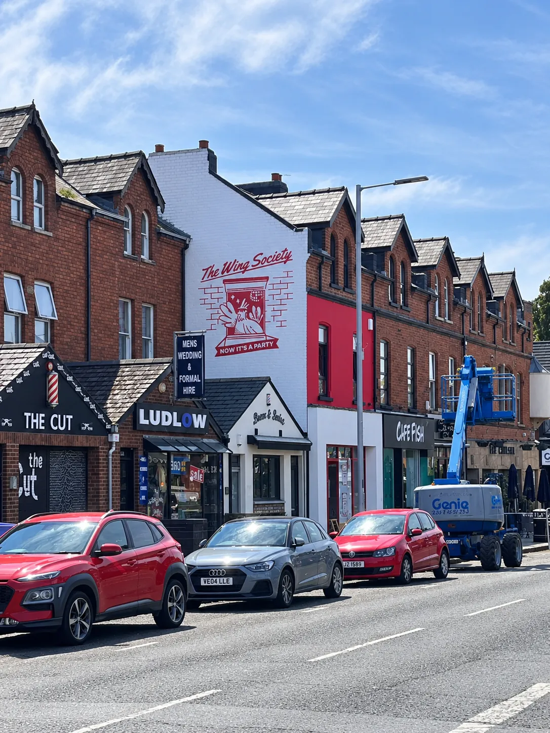 The Wing Society’s painted gable seen from down the street, above a row of shopfronts and parked cars.